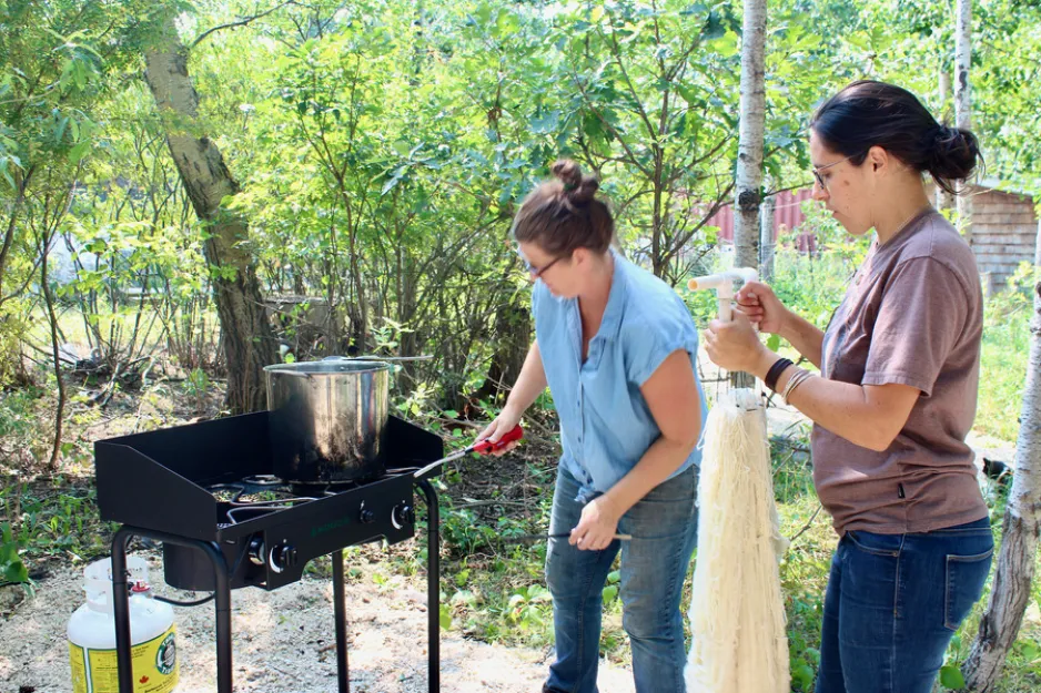 Two women stand in a sunny patch of forest. Anna Hunter, on the left, lights her outdoor stove to begin boiling water. Christel Lanthier, on the right, inspects the dyeing frame the wool will hang from. This frame is made of white PVC pipe and skeins of wool are attached to it with zip ties.