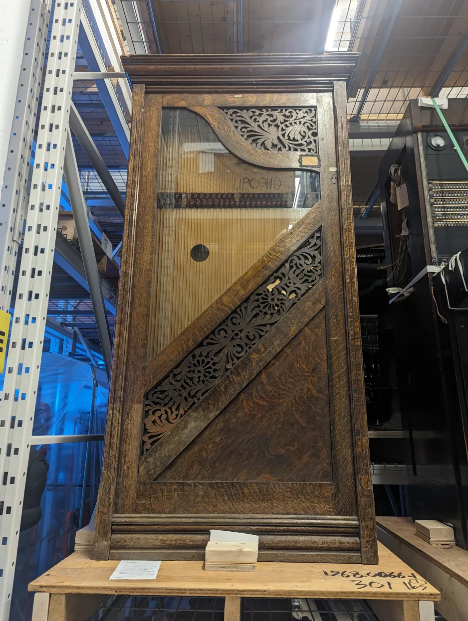 Wide, front angle shot of Whitlock Automatic Harp, sitting on a pallet in collections storage. Harp is positioned upright, approximately the height of a person. It features a wooden cabinet with trim on bottom, sides, and top. It has a front glass window, in the shape of a harp, which is surrounded by intricate carved scrollwork. Through the window, strings are visible over a soundboard.