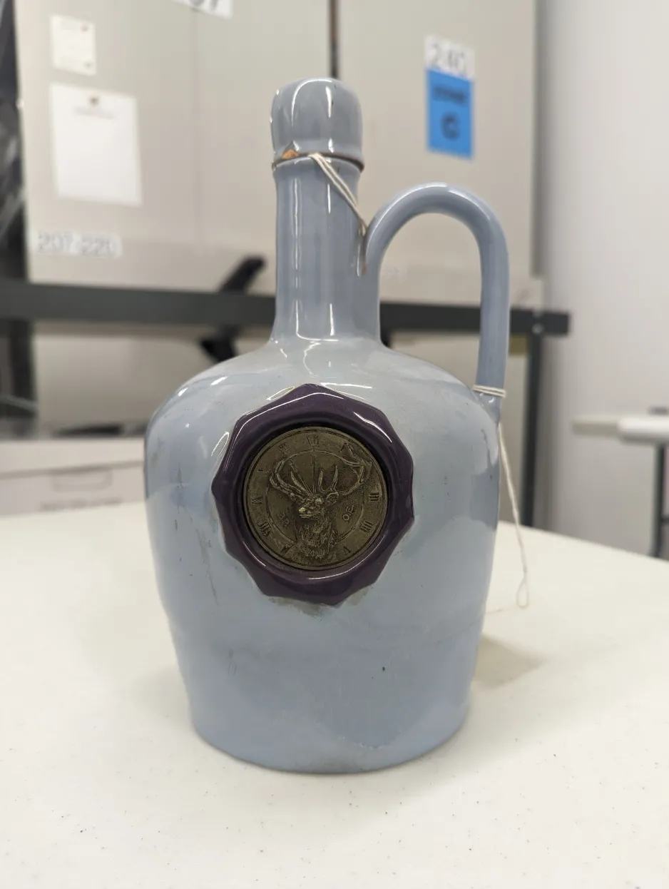 Musical jug sitting on a white table in collections storage. Jug is a powder blue decanter made of stoneware or ceramic with a glossy finish, with a high, looping handle positioned on top of the jug on one side, next to its neck. A navy-blue decagon has been sculpted into the side, which is inset with a metal medallion featuring the logo of the Benevolent Protective Order of Elks: an elk’s head in a three-quarters view, facing to the left, positioned in front of a clock face with roman numerals.