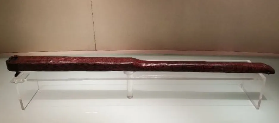 Vue d’ensemble d’un « junzhong », un petit instrument en bois présenté sur une base en plexiglas. L’instrument a un fini laqué, lisse et foncé, et a à peu près la taille et la forme d’une petite flûte, mesurant 115 centimètres de longueur.