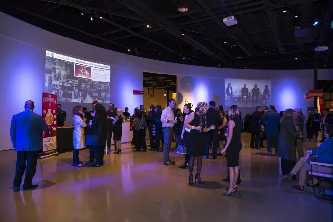 A large room in the middle of the Canada Science and Technology Museum. The walls are curved and white, the ceiling is high and painted black with cool-toned blue and white spotlights shining down. Two different images are projected onto the different places on the white wall. Many people in formalwear are socializing in small groups.