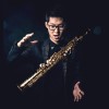 Studio photo of the author, a young Asian man with short black hair, head slightly bowed, wearing a pair of black-rimmed glasses with lips slightly open. He is wearing a black shirt and suit with metal studs embellishing the collar of the suit. Both hands are open, one above the other, and a soprano saxophone is floating between his hands. The background is black with a vague glow. The photo looks as if a magician is manipulating a floating saxophone in the air.