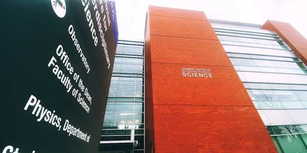 Low-angle view of signs on the University of Alberta campus. Left is a black wayfinding sign that reads “Physics, Department of” in white writing. Right is a white building sign that says “Centennial Centre for Interdisciplinary Science.” This sign is affixed to a multi-floor red brick exterior wall, with windows on either side.