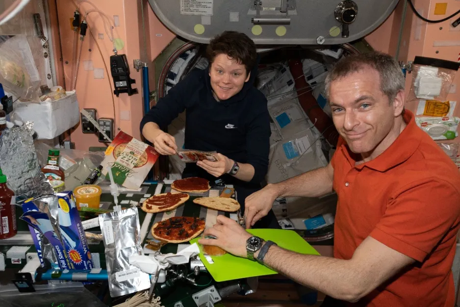 Two astronauts are smiling towards the viewer, floating before a busy countertop laced with velcro, pizza ingredients, an open cargo box, and a drinking bag. Four small, round personal-sized pizza crusts lay on the counter directly before them, partly covered in tomato sauce and black olives. Behind them is an open hatch with a room full of cargo. The peach-coloured walls are covered with wipe dispensers and small control panels.  