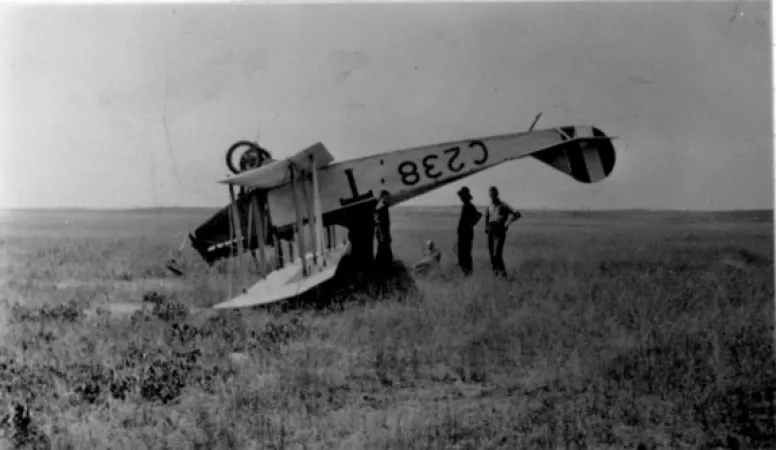 Image is a black-and-white photograph showing a Curtiss JN-4 biplane crashed in a field. The airplane is completely upside down.