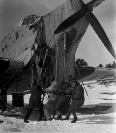 Image is a black-and-white photograph showing four men pulling on a rope attached to a pulley, with another man standing on a ladder. They are next to the nose of a large aircraft. 