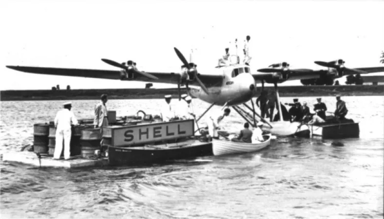 Black and white photograph showing a seaplane being refueled on the water at a dock with a Shell sign on it.  