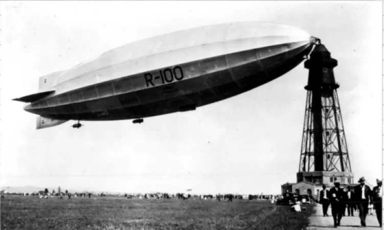 Black and white photograph of the R-100 airship docked at a mooring station tower that rises up above the people walking in the foreground.