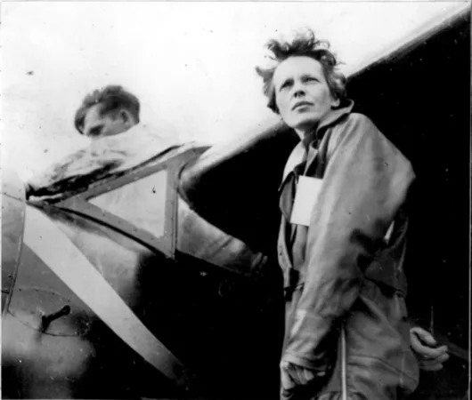 Black and white photograph of pilot Amelia Earhart standing under the wing of her aircraft as a man works on something in the cockpit.   