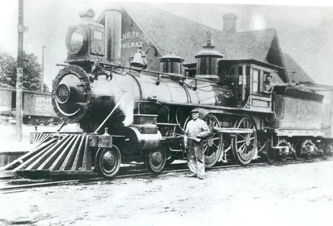 Image is a black-and-white photograph showing a steam locomotive in front of a station building. Grand Trunk is visible on the side of the station. A workman with an oil can is posing in front of wheels of the locomotive. 