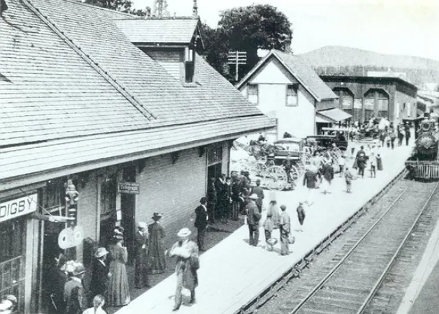 Image is a black-and-white photograph showing people waiting on the platform of the train station in Digby, Nova Scotia. A steam locomotive is approaching on the tracks on the right side of the photograph.