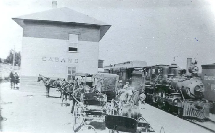 Photographie en noir et blanc montrant une locomotive à vapeur sur la voie à côté de la gare de Cabano, au Québec. Il y a des chevaux qui tirent des calèches alignées à côté de la piste et de la gare.