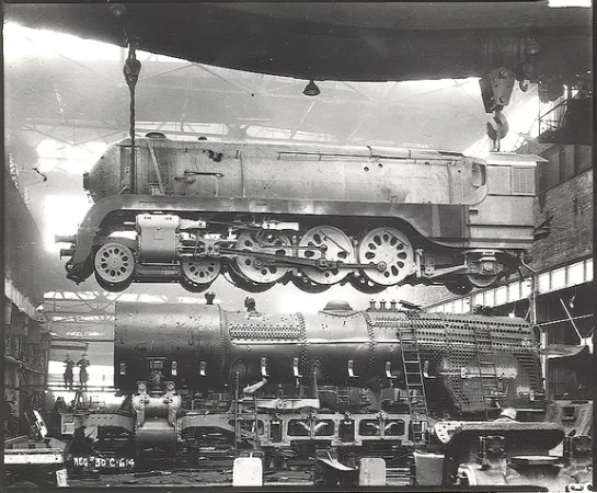 Photographie en noir et blanc d’une locomotive à vapeur en construction. Elle montre une locomotive suspendue dans les airs et des pièces sur le sol.