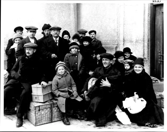 Image is a black-and-white photograph of a group of immigrants standing or sitting with luggage, perhaps on a station platform. They are dressed warmly with hats and scarves.