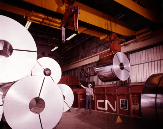 Image is a colour photograph showing a worker pointing up at coils of aluminum that is being unloaded in a CN yard. 