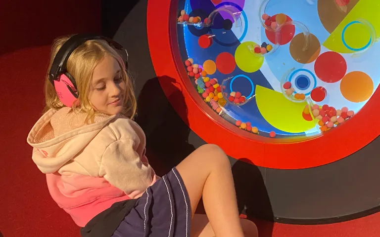Young child in a skirt and a pink hoodie wearing pink noise cancelling headphones is sitting inside a colourful circular interactive. 