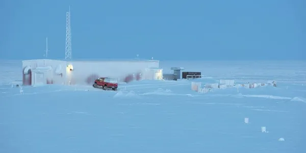 A barren, snowy landscape is set against a blue sky. A research facility for Environment and Climate Change Canada can be seen, with its lights glowing.