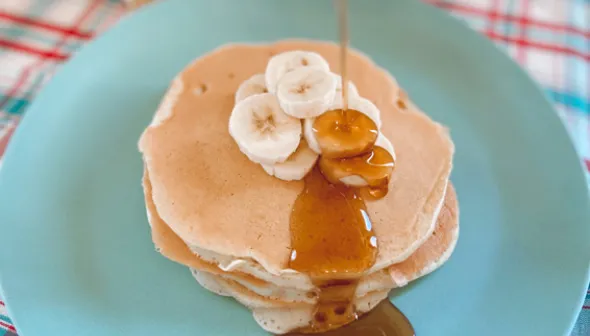Maple syrup pours out of a glass jar and on to a stack of pancakes and sliced banana below. The food sits on a blue plate, on top of a plaid tablecloth.