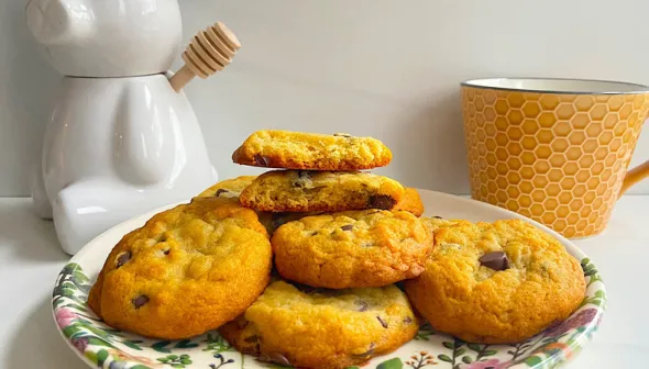 A pile of chocolate chip cookies sit on a colourful plate. A bear-shaped honey container and a mug with a honeycomb design are visible in the background.