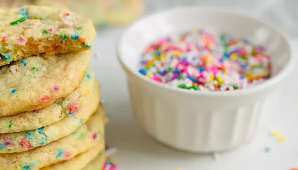 A cookie sits on a white surface. Next to it, a stack of cookies and a bowl filled with sprinkles.