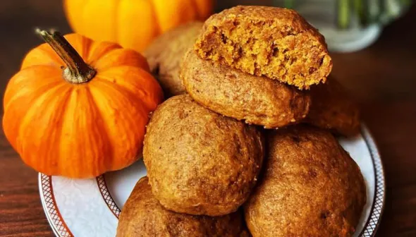 A small pile of brown-coloured pumpkin cookies sit on a plate, next to some small orange gourds.