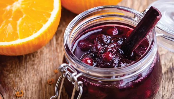 A glass jar filled with cranberry sauce sits on a wooden surface. Orange slices are visible in the background.