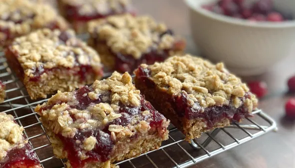 Rows of cranberry oatmeal bars sit on a metal baking rack; a bowl of cranberries is visible in the background.