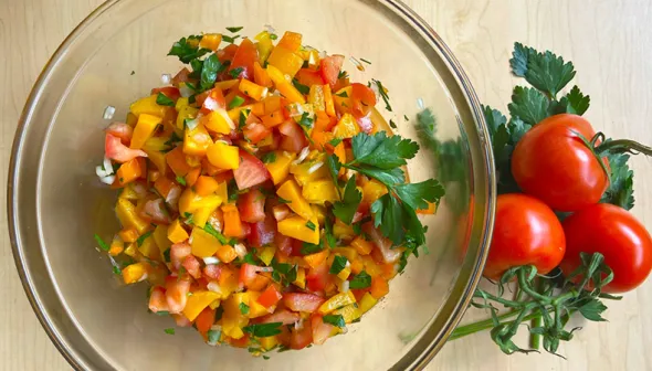 An aerial view shows a clear bowl filled with fresh salsa, set against a light tabletop. Three ripe tomatoes sit next to the bowl.
