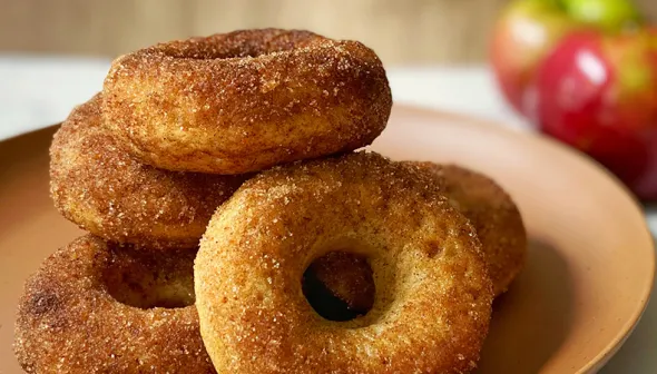 A plate is piled high with donuts; apples are visible in the background.