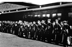 L’image est une photo noir et blanc montrant le premier contingent de soldats canadiens au garde-à-vous devant une voiture de train. 