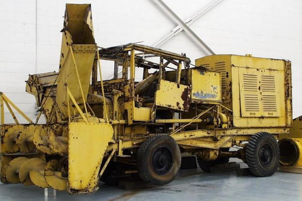 A large yellow snowplow with various rusting spots sitting on the floor of a warehouse.