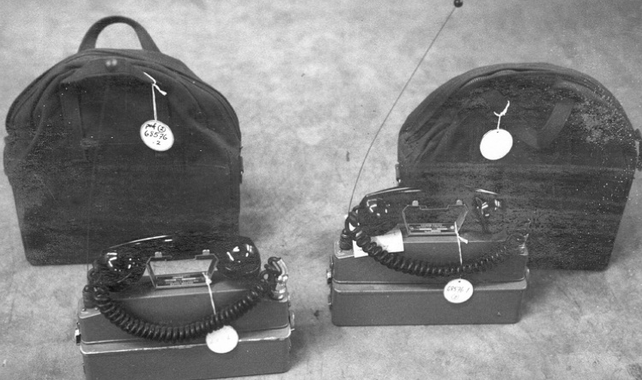 Une photo en noir et blanc de deux radios émettrices-réceptrices placées devant leurs sacs de rangement. 