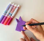 La main d'un artiste colorie la forme du lapin en origami sur une surface blanche.   Quatre marqueurs de couleur se trouvent également sur la surface blanche.