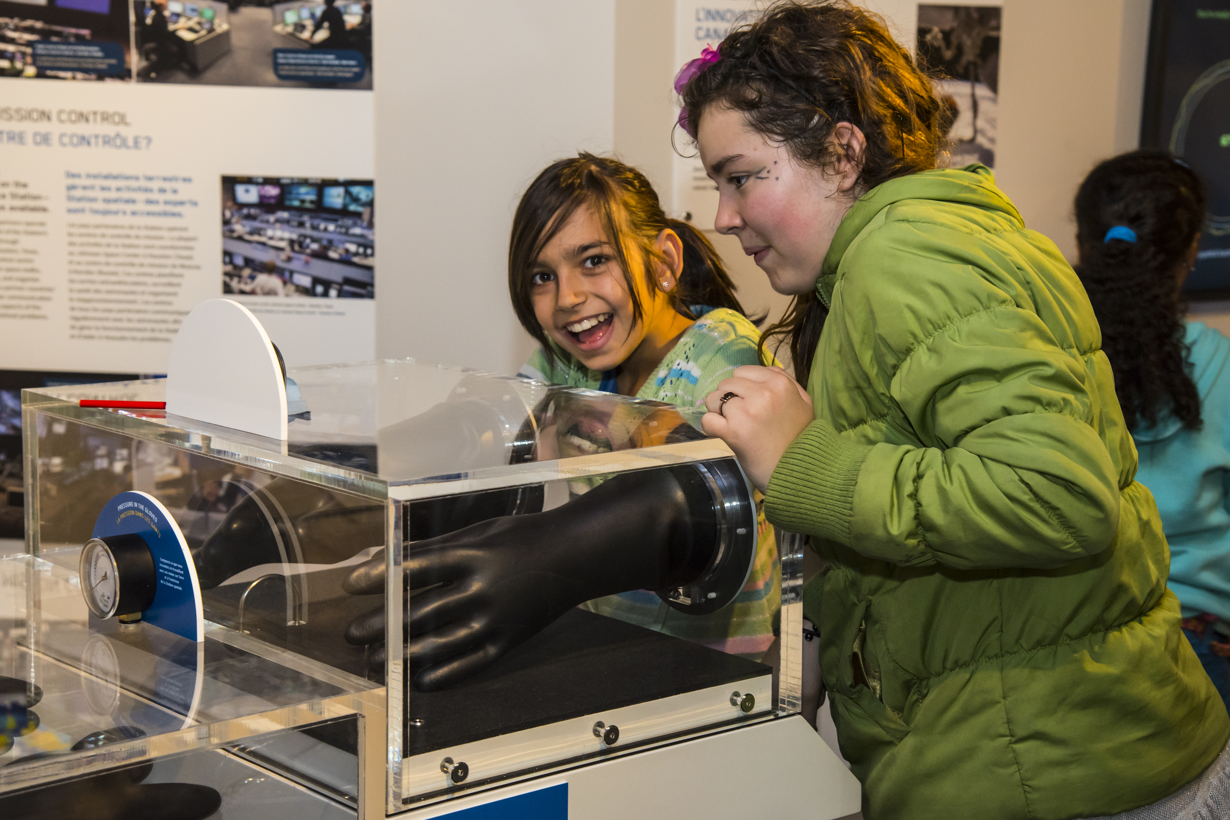 Two young girls stand next to a glass case in a museum; one girl is inserting her hand into a black pressurized glove inside the case.