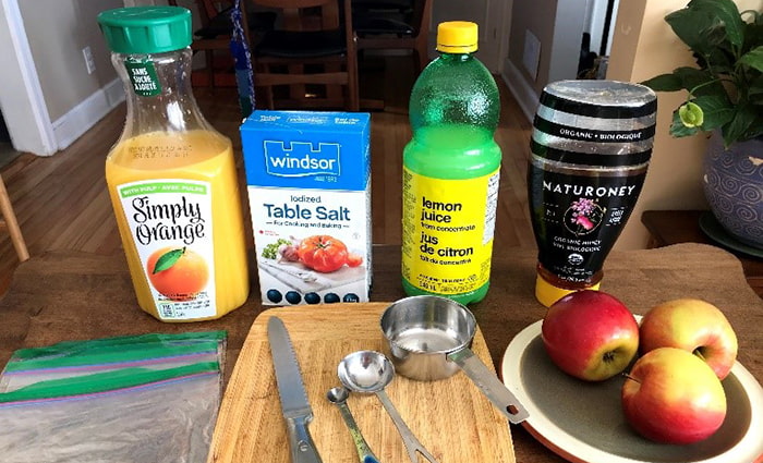 Les ingrédients sont disposés sur une table avec une planche à découper et des ustensiles. À l’arrière, on voit un pichet de jus d’orange, une boîte de sel de table, une bouteille de jus de citron et une bouteille souple de miel ainsi que trois pommes rouges et jaunes dans une assiette sur le côté.