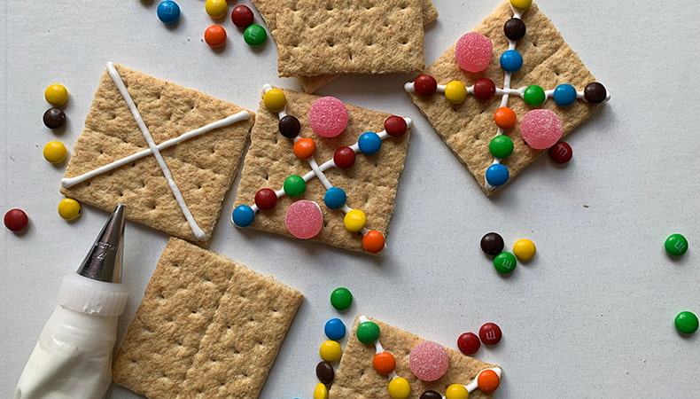 Graham crackers decorated with icing and colourful candies sit on a white surface, along with a piping bag with metal tip.