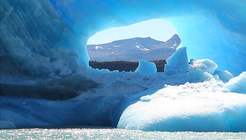 Un énorme morceau de glace flottant dans l'eau remplit le cadre.  Un trou au centre de la glace nous permet de voir une montagne au loin derrière.