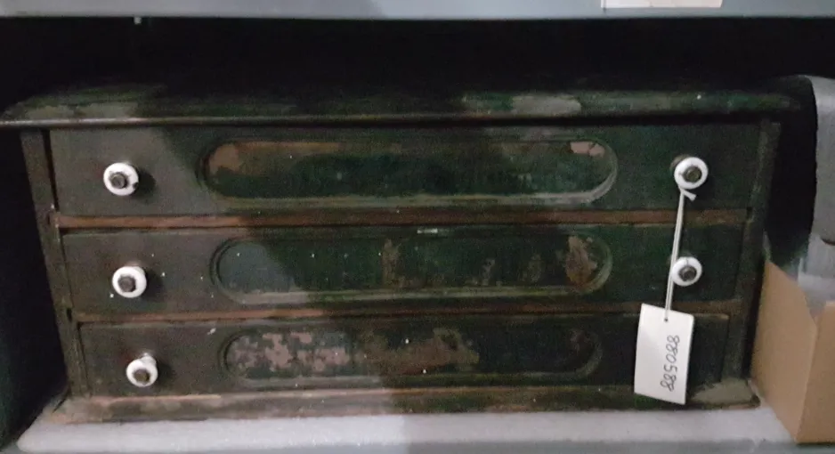 A wooden cabinet with three drawers sits on a grey shelf. There is dark paint peeling on the front, and each drawer has two circular, white drawer pulls. One drawer pull is not visible as it is covered by a white tag that reads “880588” in black.