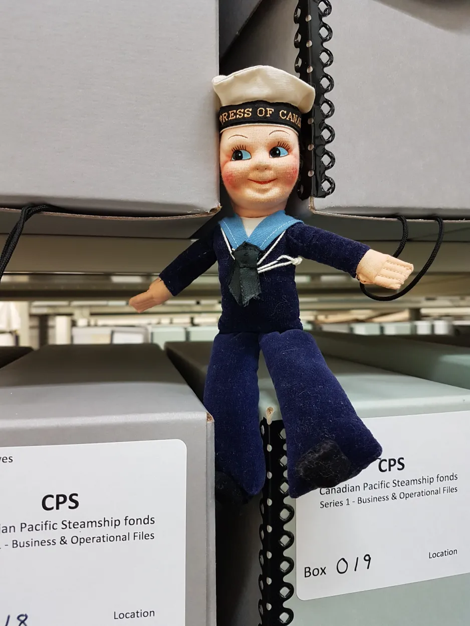 Photograph of a sailor doll sitting on a shelf in the archives between archival boxes.