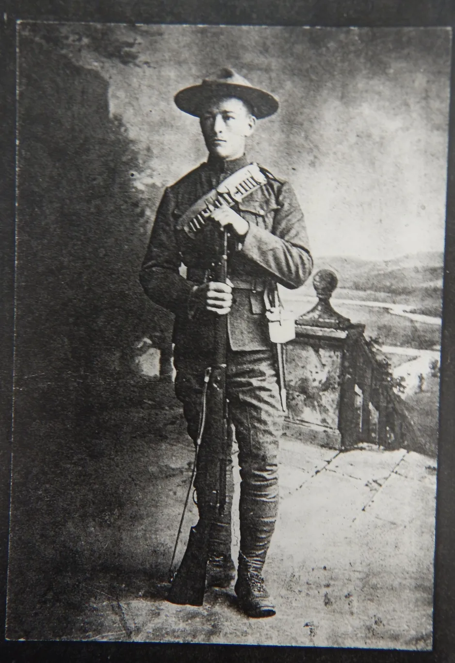 Photographie noir et blanc montrant un homme en tenue militaire faisant la pose avec un fusil. 