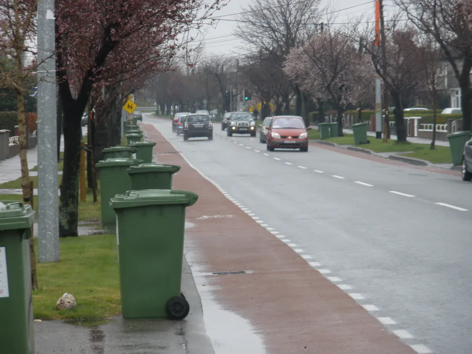 Rows of green bins line a rainy residential street, awaiting collection.