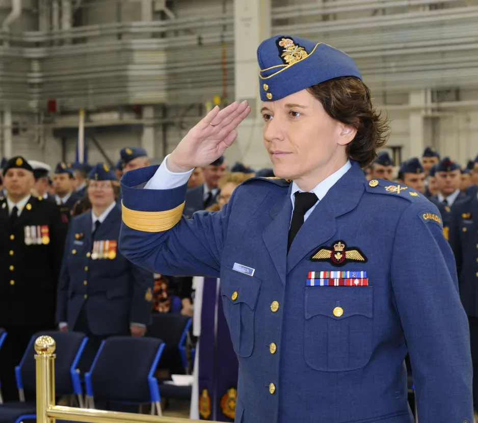 A woman in a blue military uniform stands at attention and salutes in front of a crowd of people.