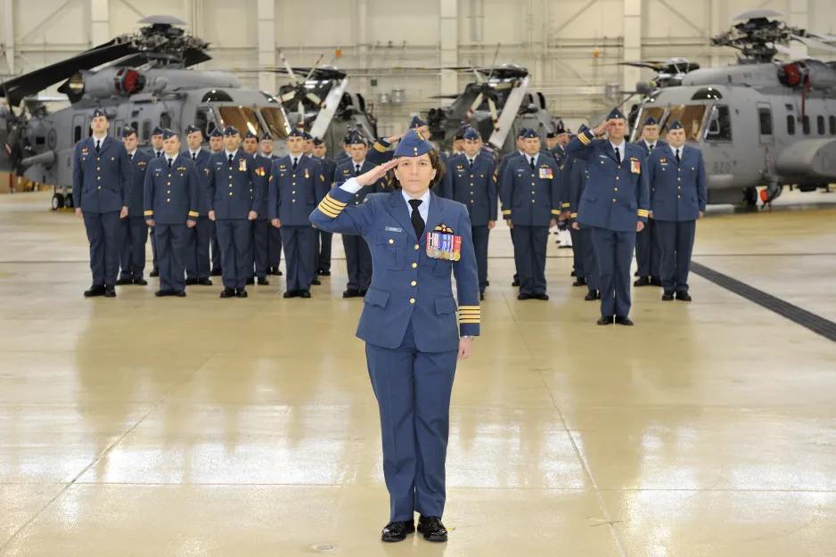 A woman in a blue military uniform stands at attention and salutes, in front of a squadron of men in uniform.