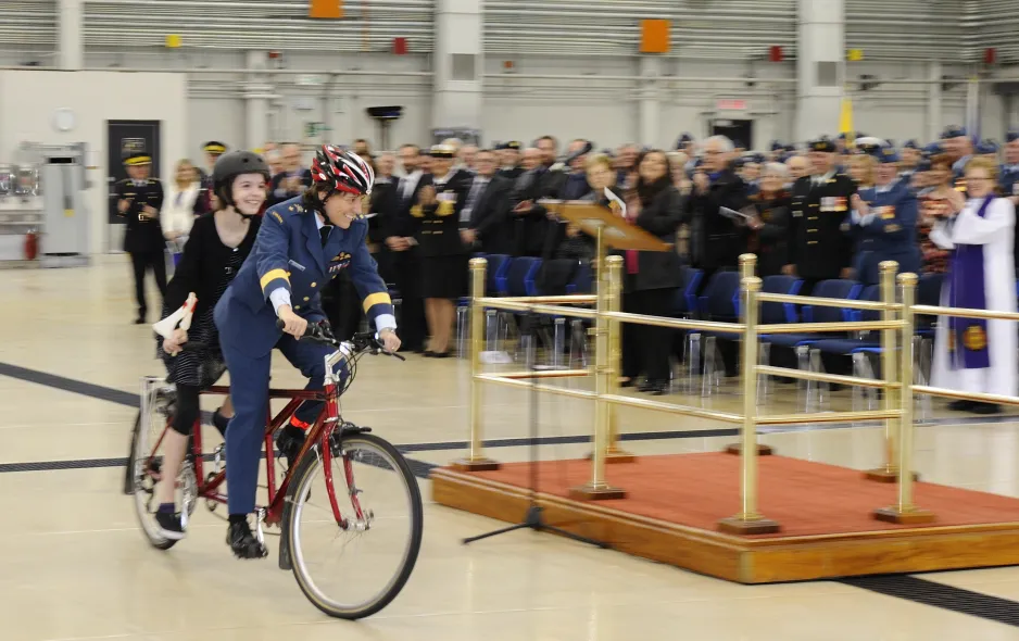 A woman in a blue military uniform rides a tandem bike with a young girl, at a ceremony attended by a large crowd.