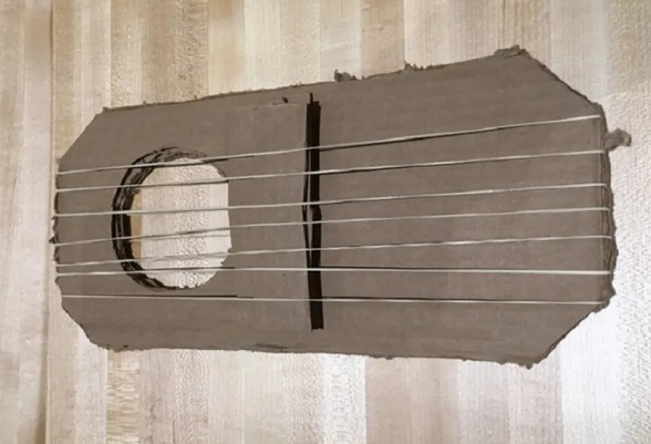 Several elastics are stretched over layered pieces of cardboard, with a hole cut through the stack to make a neckless guitar. 