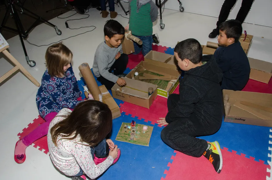 Six children sit on the floor, creating small games out of cardboard boxes.