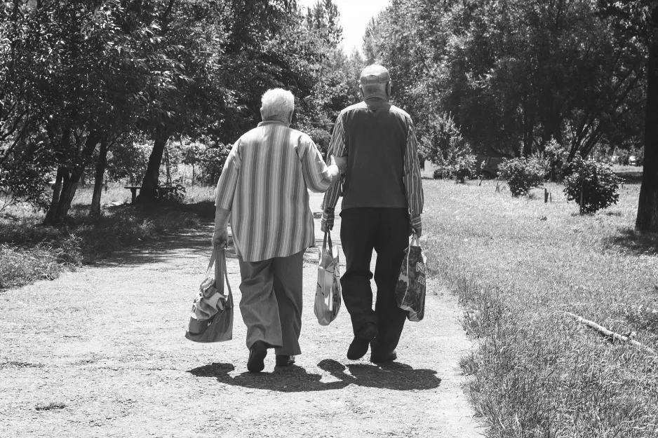 An elderly couple strolls down a treed lane carrying their groceries