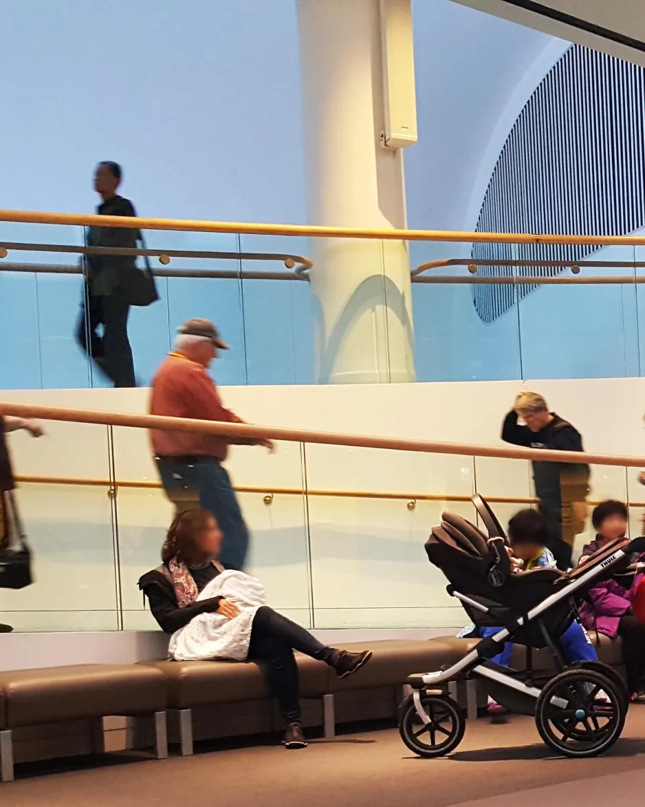 A young woman sits on a bench inside a busy museum, breastfeeding a baby draped with a blanket.