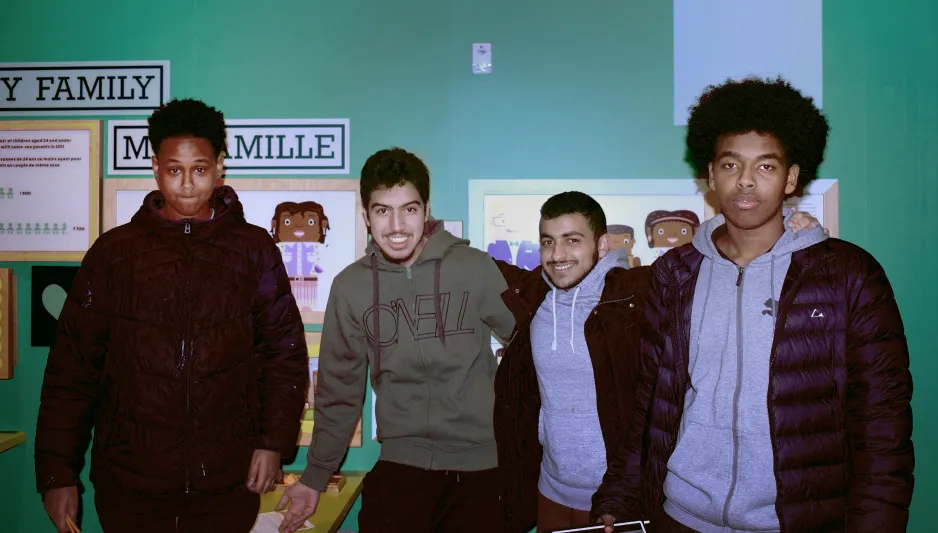 Four teenage boys pose in front of the posters and words across the wall saying my family. On the left the teenager wore a dark blue jacket, next to him the other boy wore a green sweater, followed by another teenager wearing a grey hoodie with a black jack around it and on the far right the last teenager wore a grey hoodie with a dark blue jacket overtop. 