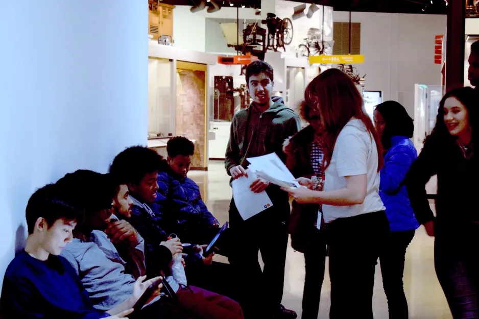 Five teenage boys sit across a bench in the museum. Standing above them is three teenage girls and a teenage boy, along with the project coordinator. She is holding out papers with instructions on them for the scavenger hunt game they were playing. 